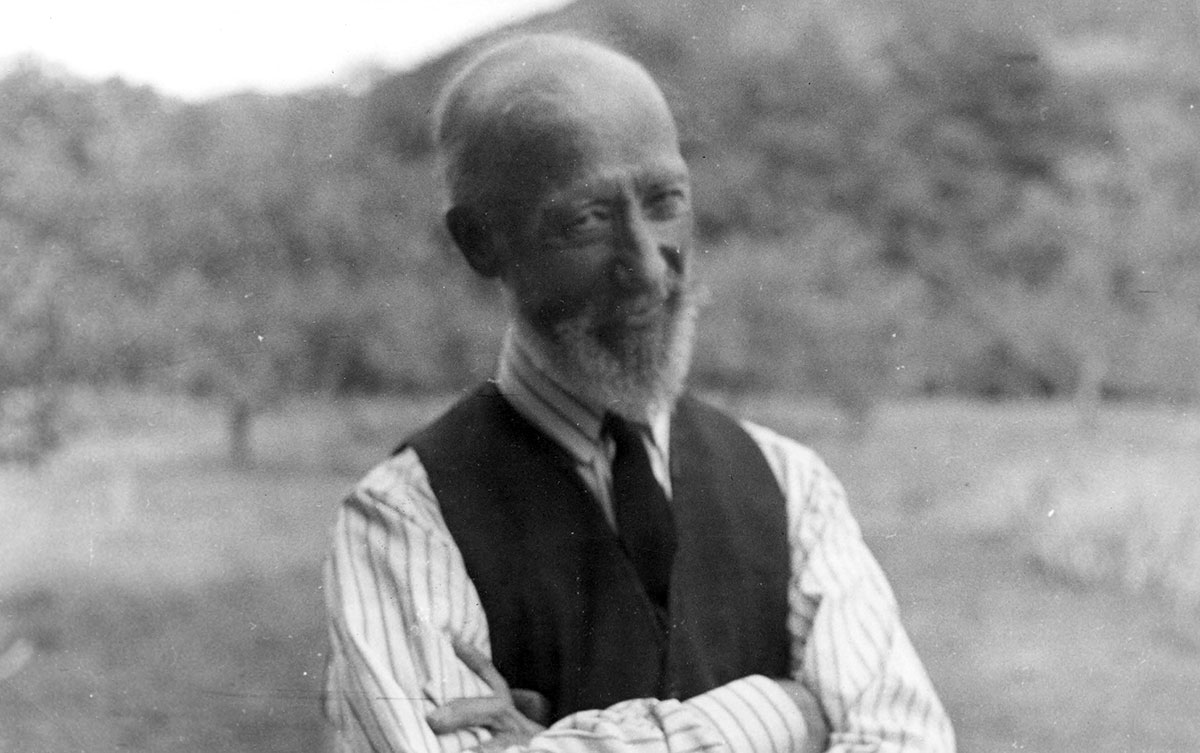 Black and white photograph of Ozias Leduc, waist up. He is bald, has a beard, and wears a striped shirt, waistcoat and tie, with his arms crossed. He looks at the camera, with a smile and a friendly gaze. Behind him we can make out an orchard and a mountain.