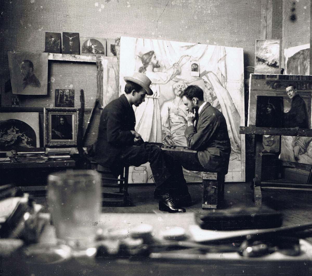 Ozias Leduc et son frère se font face, assis, concentrés devant une partie de dames. Ils sont dans l'atelier de l'artiste, avec, en arrière fond, un tableau religieux en progrès.