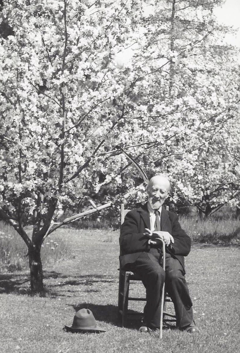 Photographie d’Ozias Leduc, assis sur une chaise, tenant sa canne entre ses jambes, dans son verger. Derrière lui se trouve un grand pommier en fleurs.