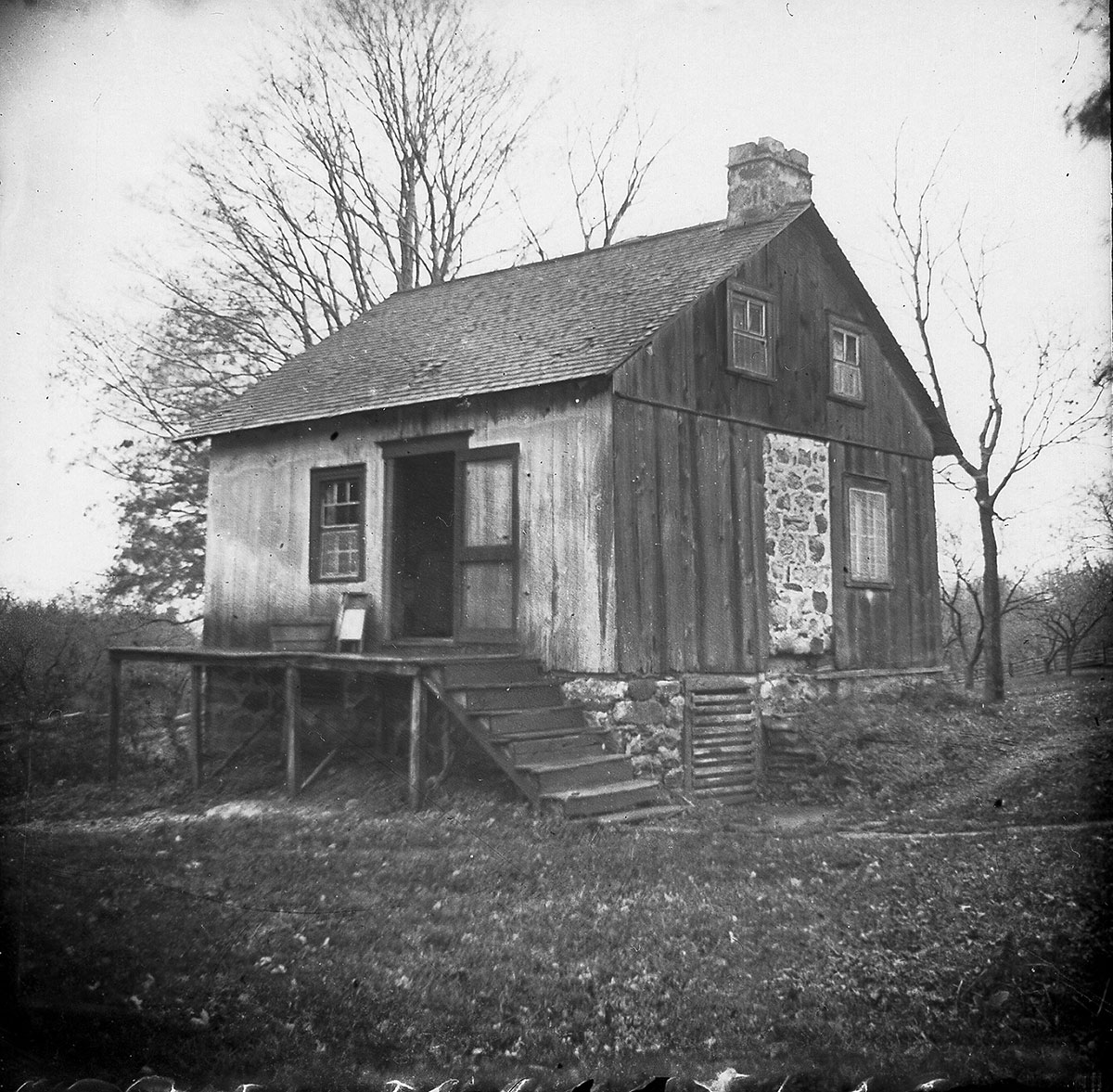 Photo illustrant une petite maison de bois, avec d'un côté une cheminée et de l'autre un escalier menant à la porte d'entrée.