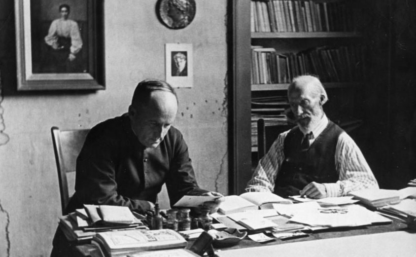 Photo de deux hommes assis derrière un bureau, Monseigneur Tessier lit un document pendant que l'artiste le regarde. Sur le mur derrière on voit une bibliothèque de même que des oeuvres de Leduc.
