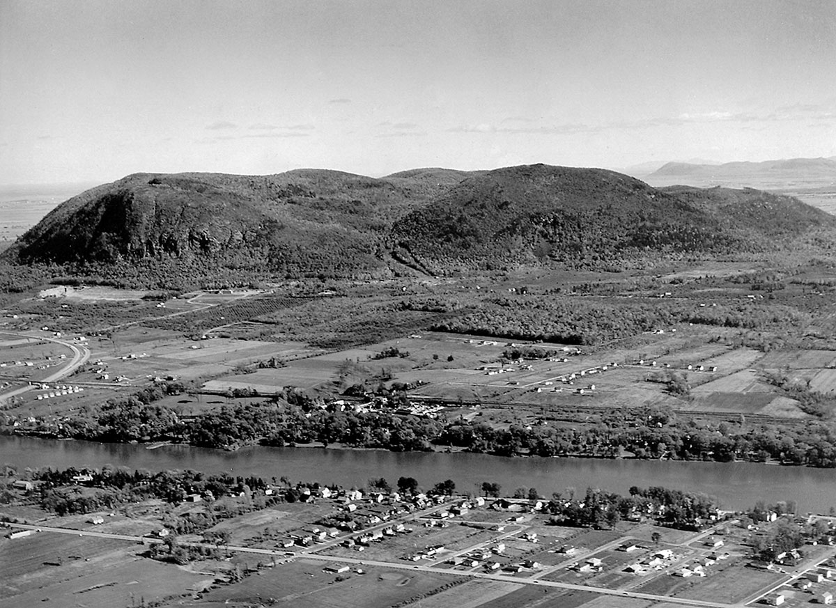 Une vue aérienne de la ville de Saint-Hilaire, en noir et blanc datant de 1955, montre la montagne et la rivière du Richelieu, les champs et les quelques maisons aux alentours.