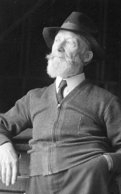 Black and white photograph of Ozias Leduc, waist up, aged 70 to 75. He wears a hat, turns his head to the right and has a beard. He has his left hand in his trouser pocket.