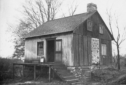 Photo showing a small wooden house, with a fireplace on one side and a staircase leading to the front door on the other.